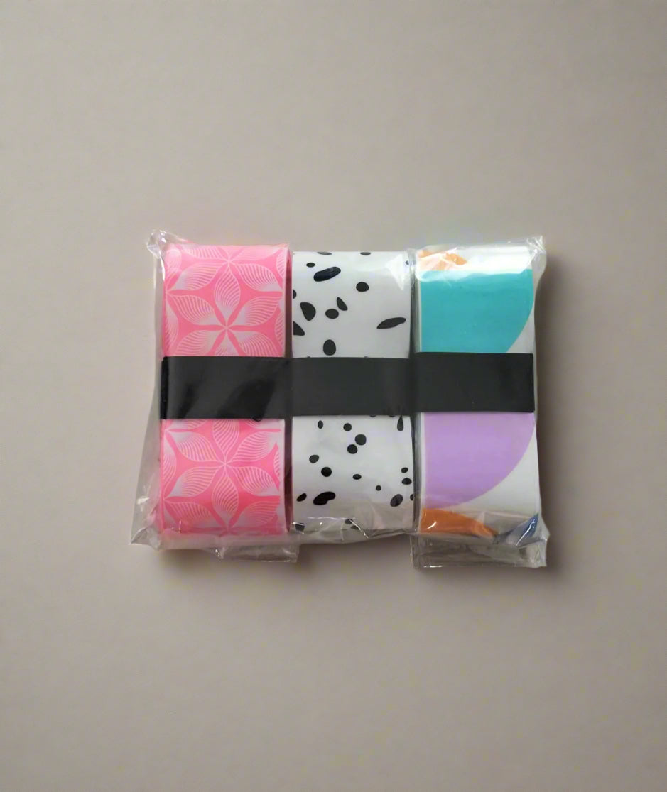 Set of 3 Pickleball grip tapes in a plastic wrapper. One of the grip tape is a pink flower design, another is black and white, like a cow design. The last is different color shapes mixed with a white background.