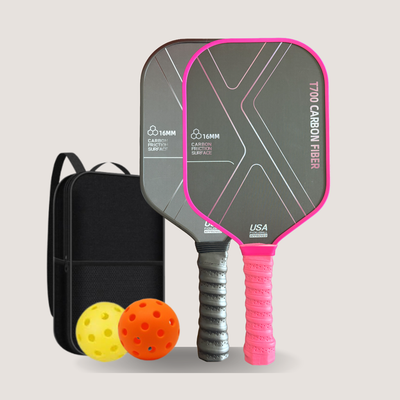 Two pickleball paddles with dark gray and pink and black handles, shown alongside a black carrying case and two balls, one yellow and one orange, on a light gray background.